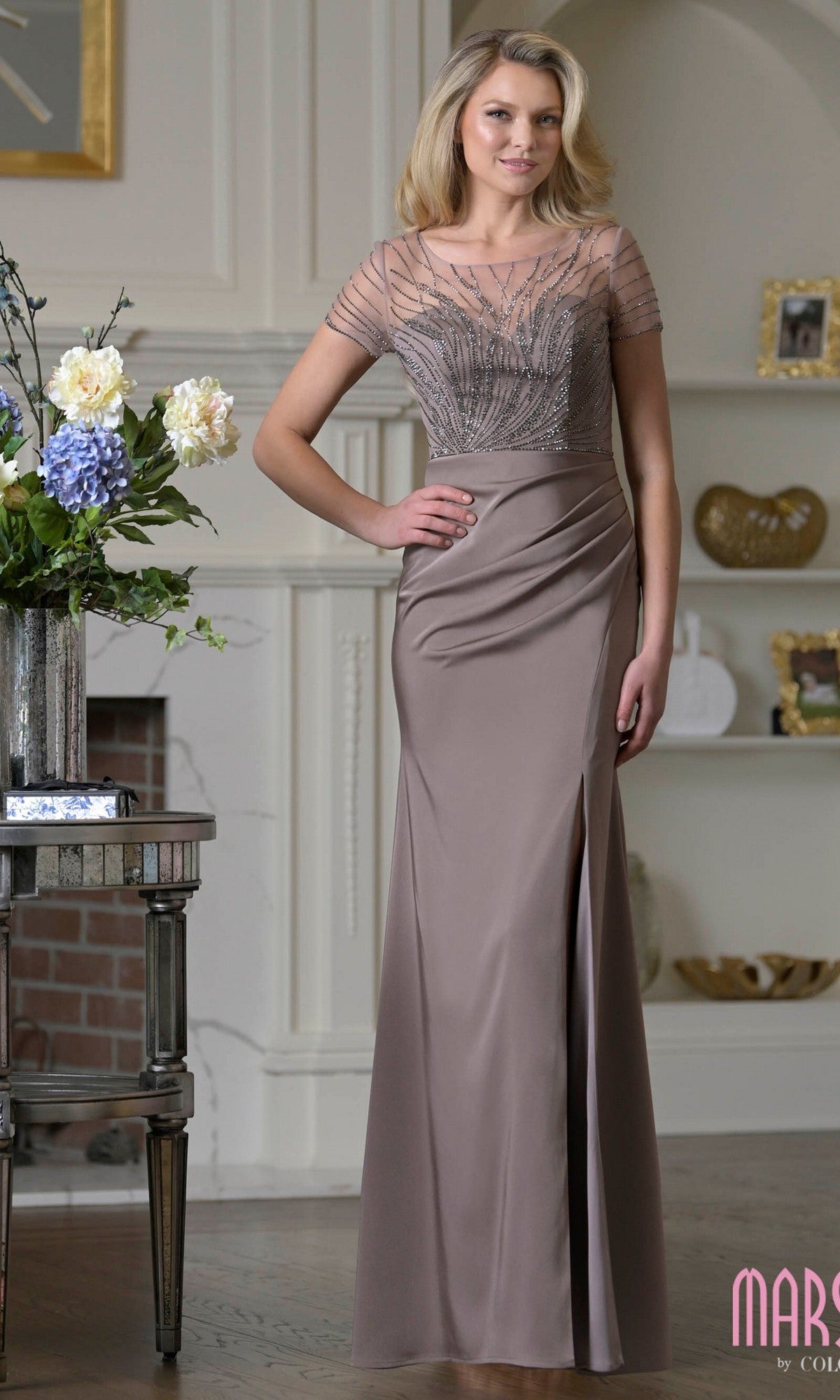 A woman stands indoors beside a floral arrangement, wearing the Marsoni M350 long formal dress in dark taupe with a sheer beaded bodice and short sleeves.